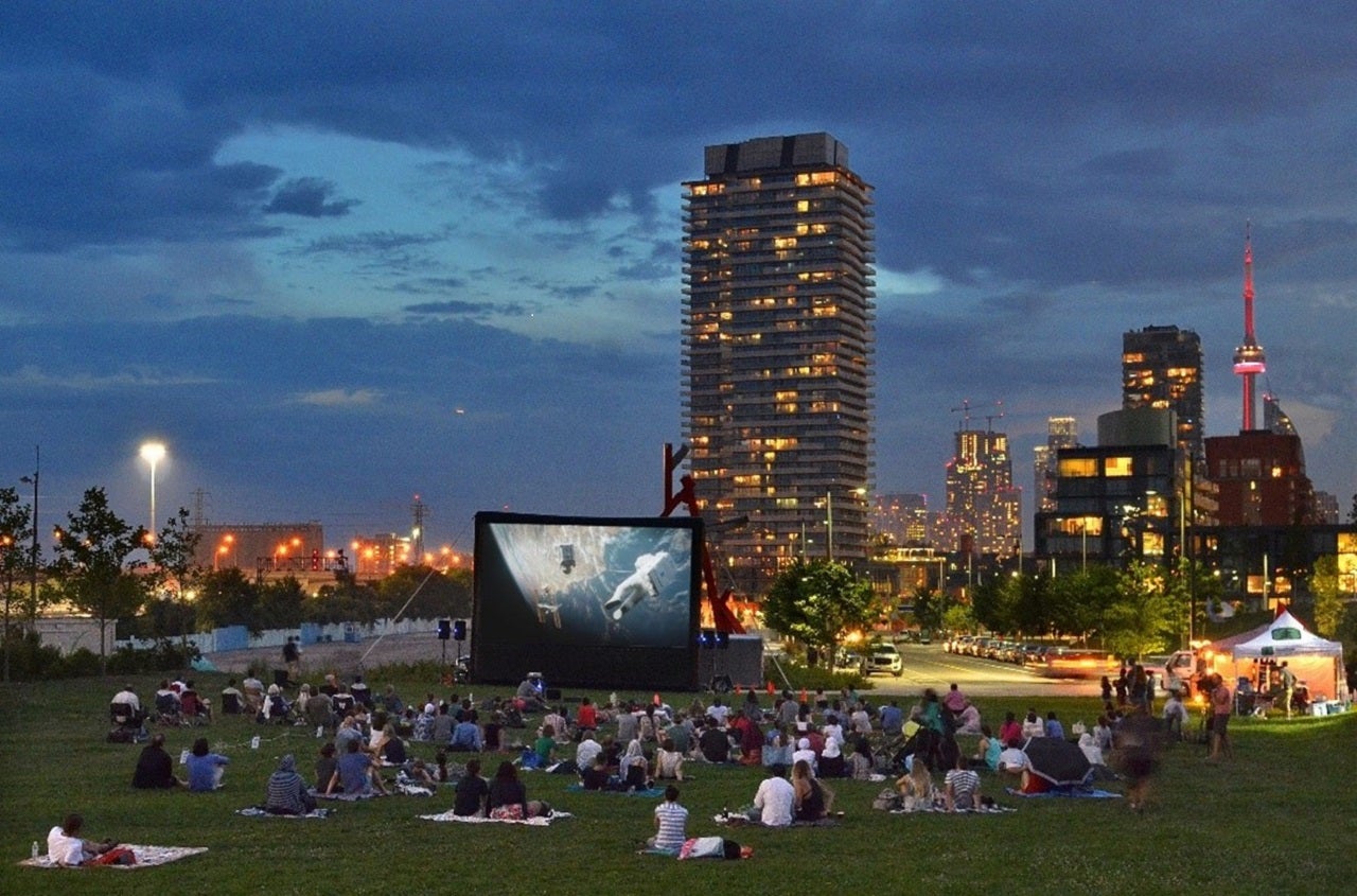 Photograph of a park with skyline and people sitting on the lawn and looking at a large screen.