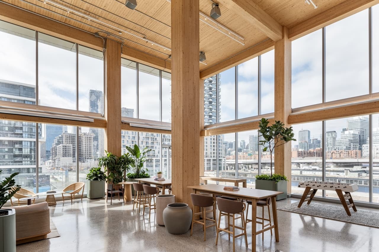 Modern office space featuring large windows with cityscape views, wooden beams, and furnishings, including tables and chairs. The interior is spacious and well-lit, with potted plants adding a touch of greenery.