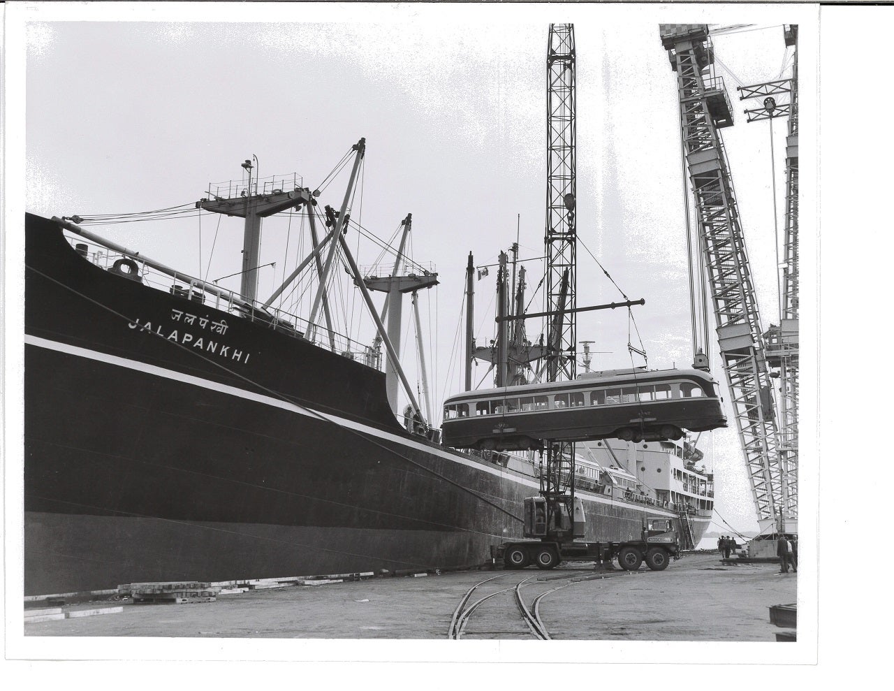 A black and white image of a ship with crane unloading a streetcar on the right. 