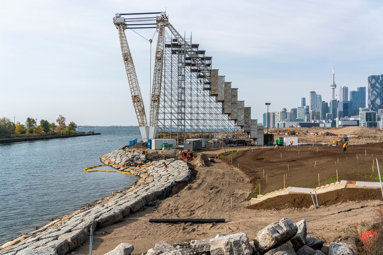 A massive crane with scaffolding on right stands tall on the edge of river amidst a construction site. 