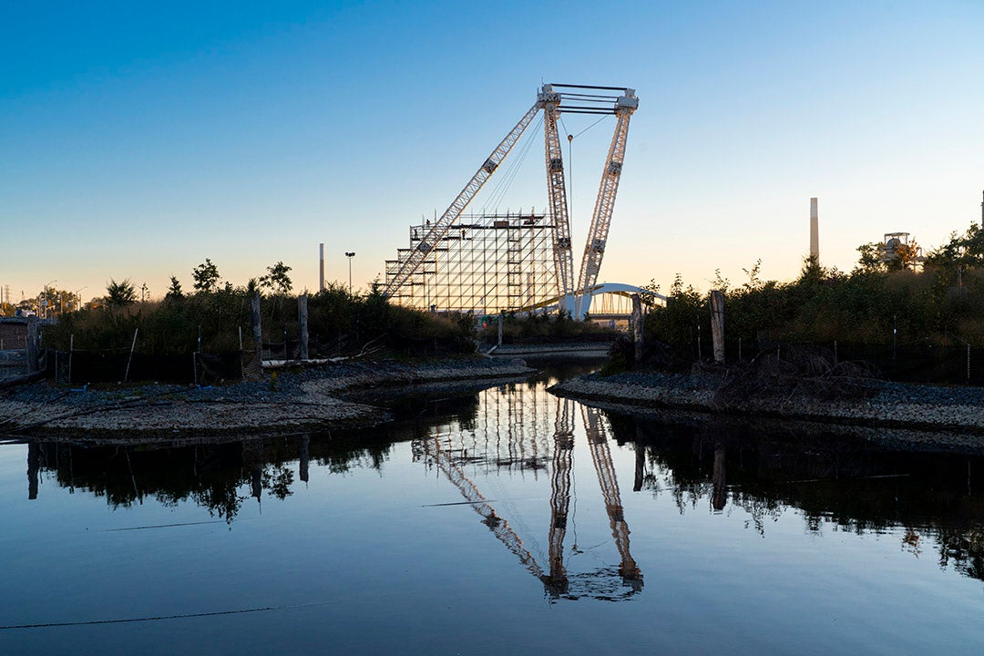 A crane standing tall on the edge of river. Its shadow being reflected in the water down. 