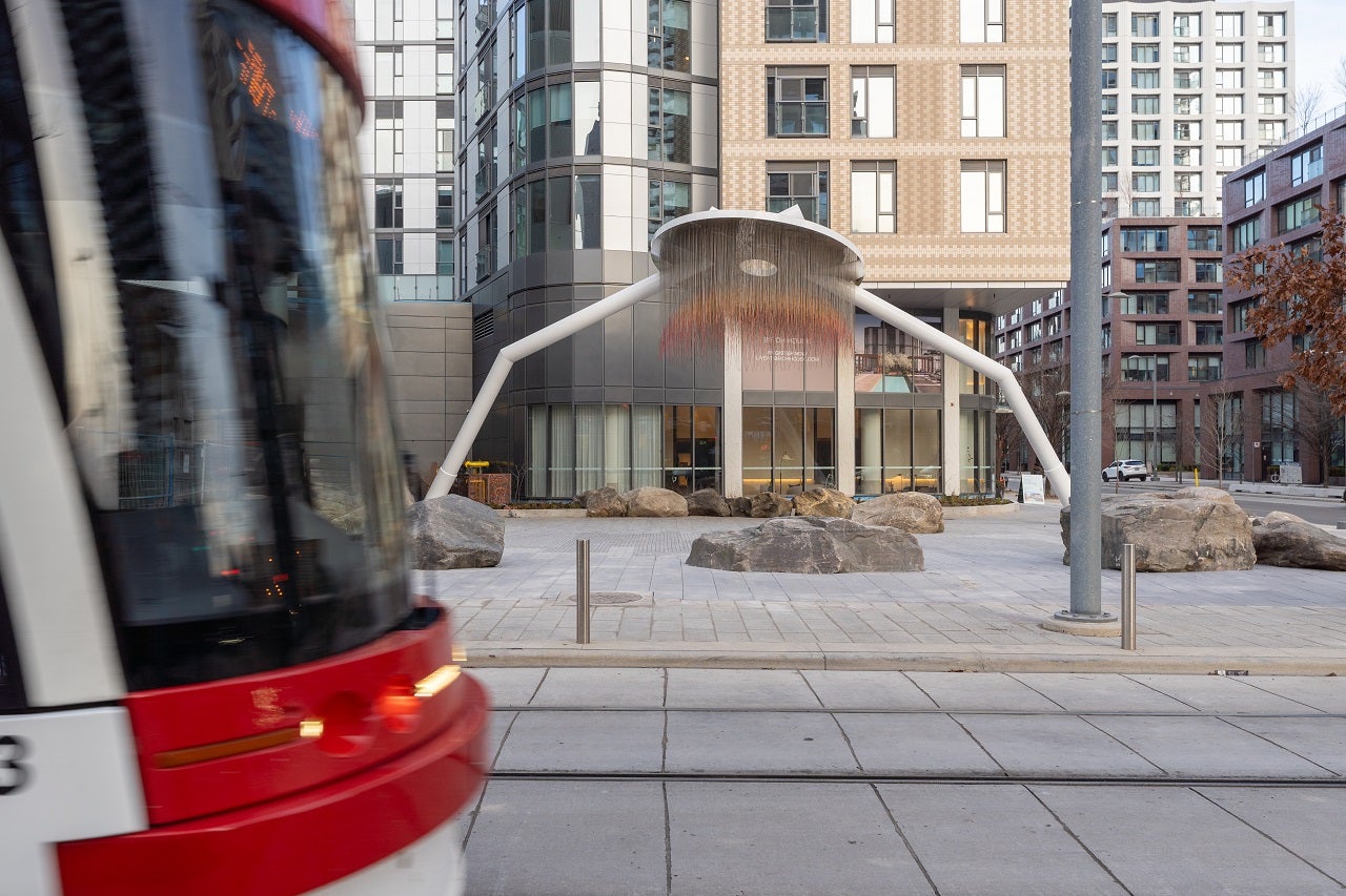 A distant photograph of a two legged art piece in front of a building with street car coming into the frame from the left.