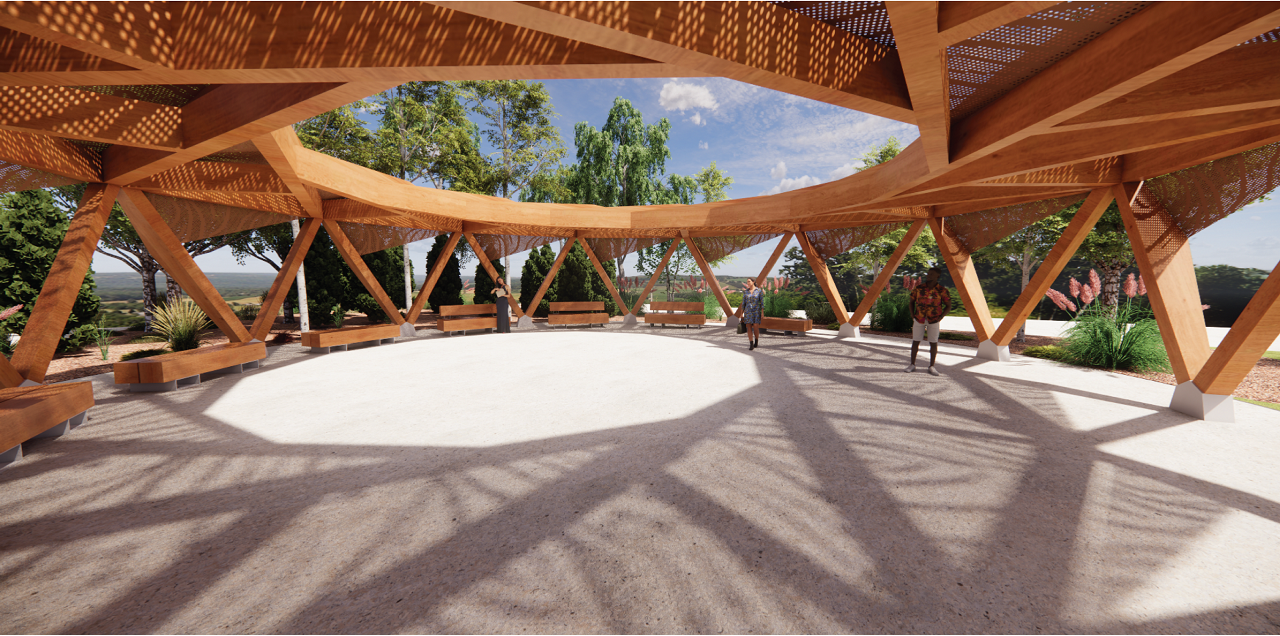 Wide view of the shade structure with people enjoying the shaded area on a sunny day, surrounded by lush greenery in the background.