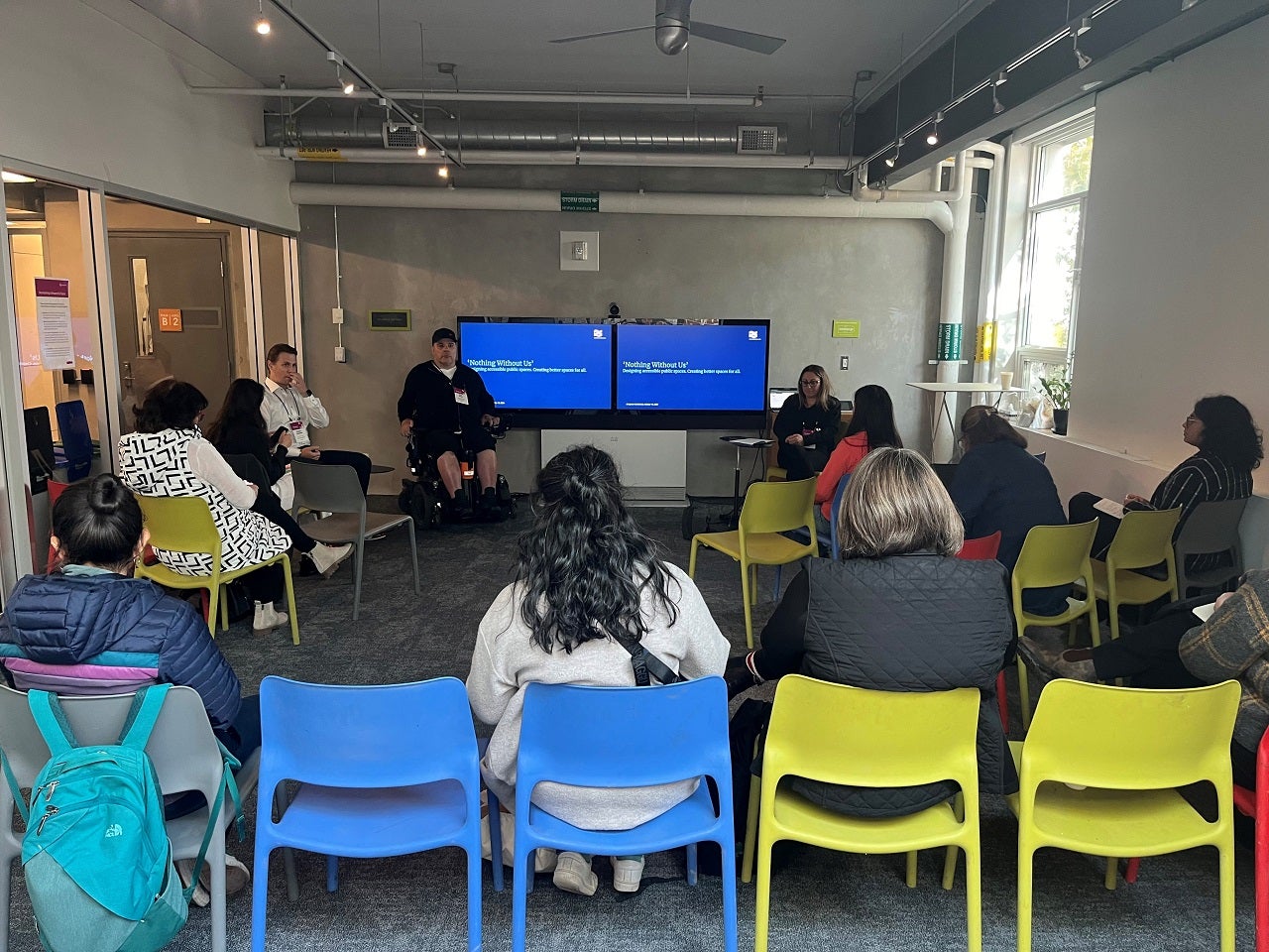 A group of individuals seated on different colours of chairs in a seminar room, attentively facing a speaker next to a large presentation screen displaying a blue slide with text.