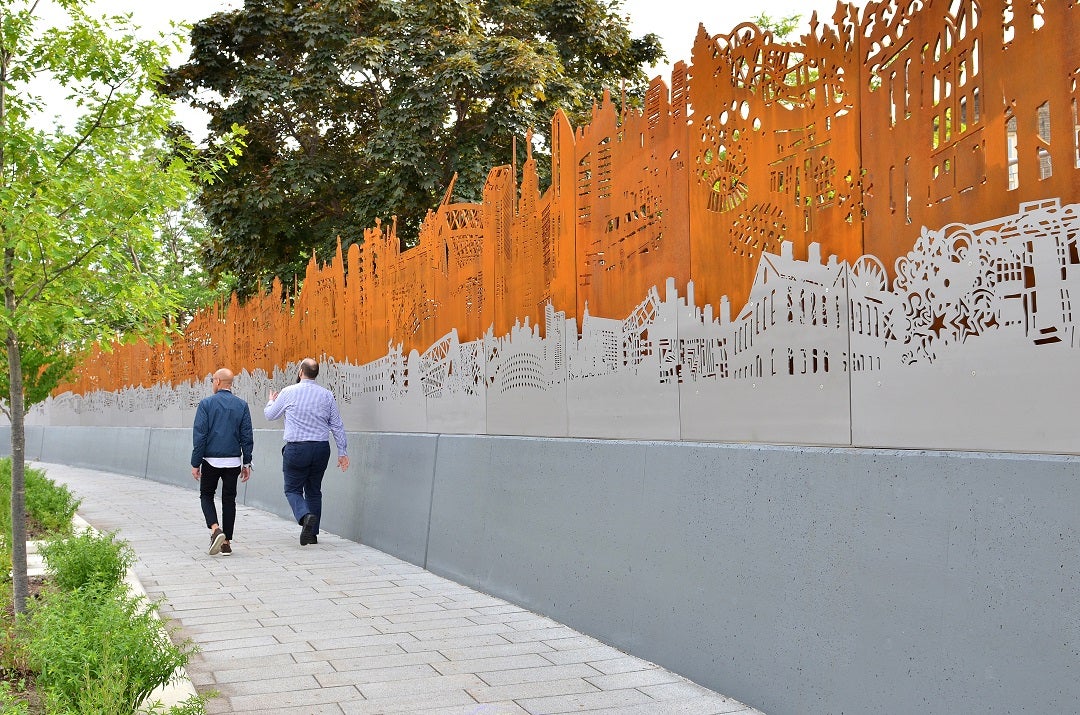 Two people walking on a sidewalk past a large wall of public art pieces