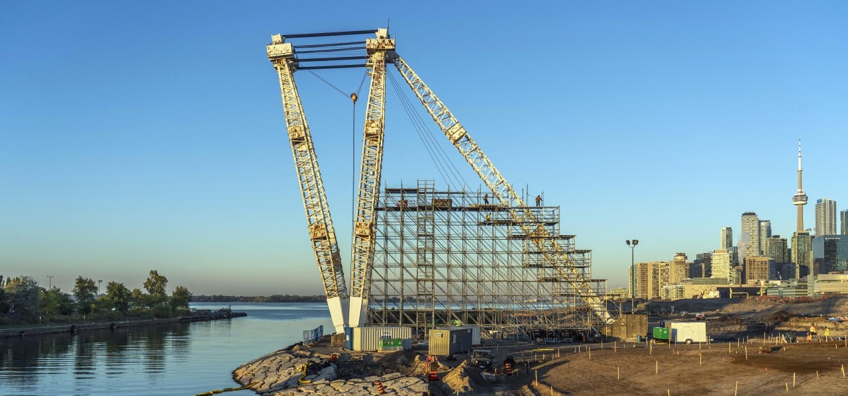 A massive crane with scaffolding stands tall on the edge of river in the middle of a construction site. 