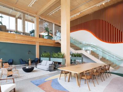 Modern office lobby with wooden accents and a variety of seating areas, featuring a large wooden table, sofas, and armchairs. The space includes lush green plants and a staircase with glass railings leading to an upper level. Decorative elements include a hanging wooden slat feature and green wall panels.
