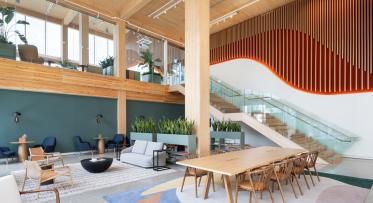 Modern office lobby with wooden accents and a variety of seating areas, featuring a large wooden table, sofas, and armchairs. The space includes lush green plants and a staircase with glass railings leading to an upper level. Decorative elements include a hanging wooden slat feature and green wall panels.